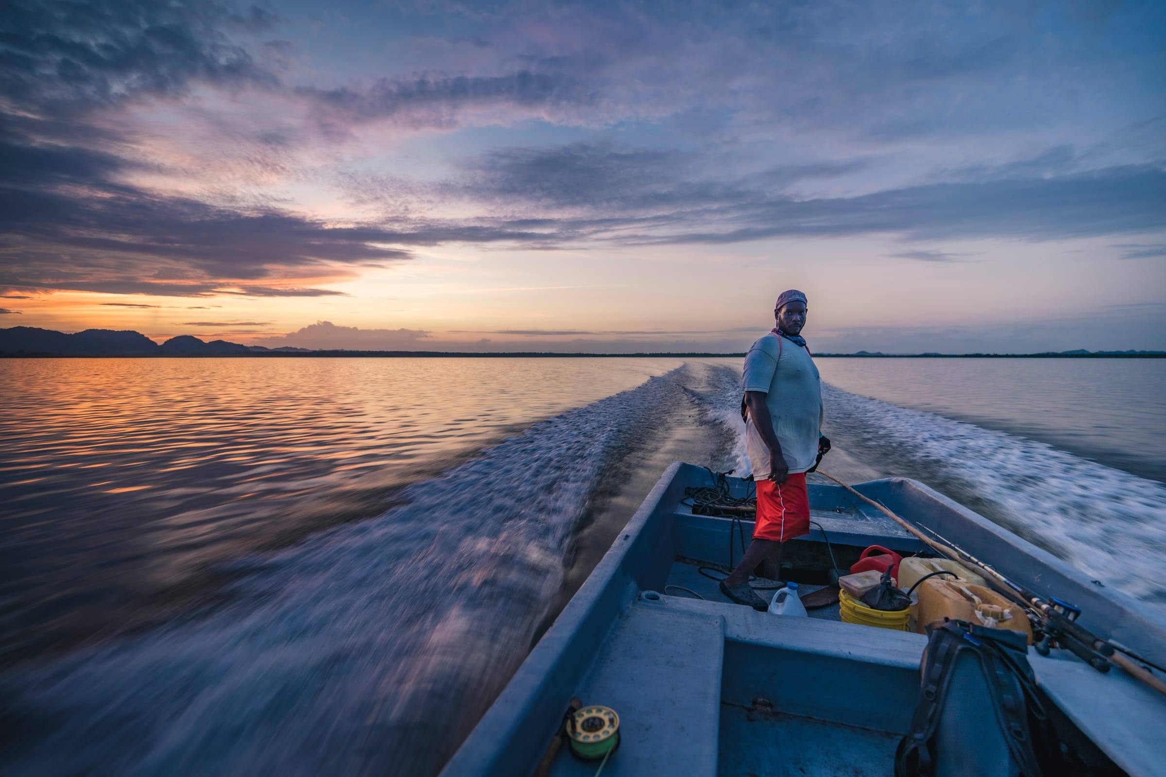 A boat captain stands at the helm of a skiff crossing a lagoon at dawn