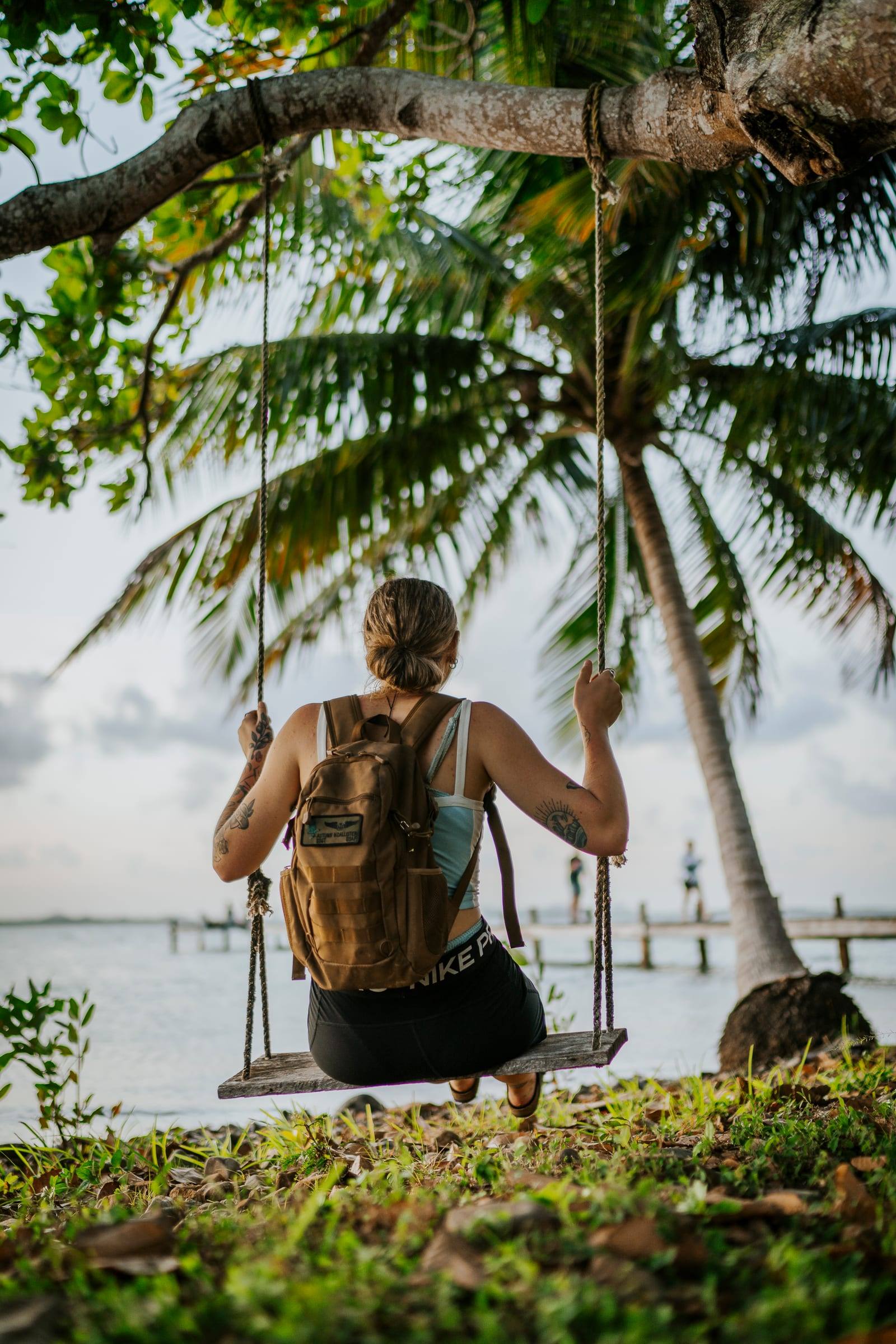 A traveler with a backpack sits on a rope swing under palm trees